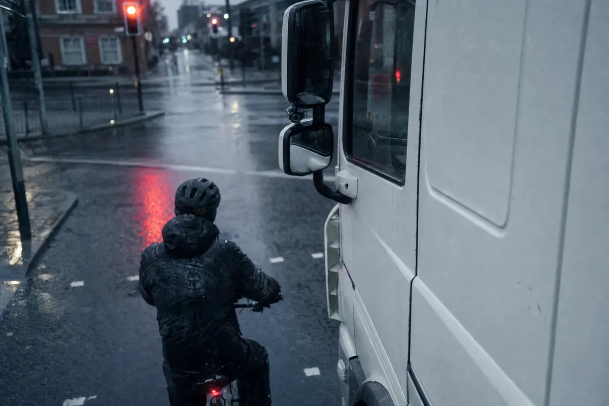 Illustrative image of a cyclist waiting beside an HGV cab at a wet city junction, with a camera on the mirror arm