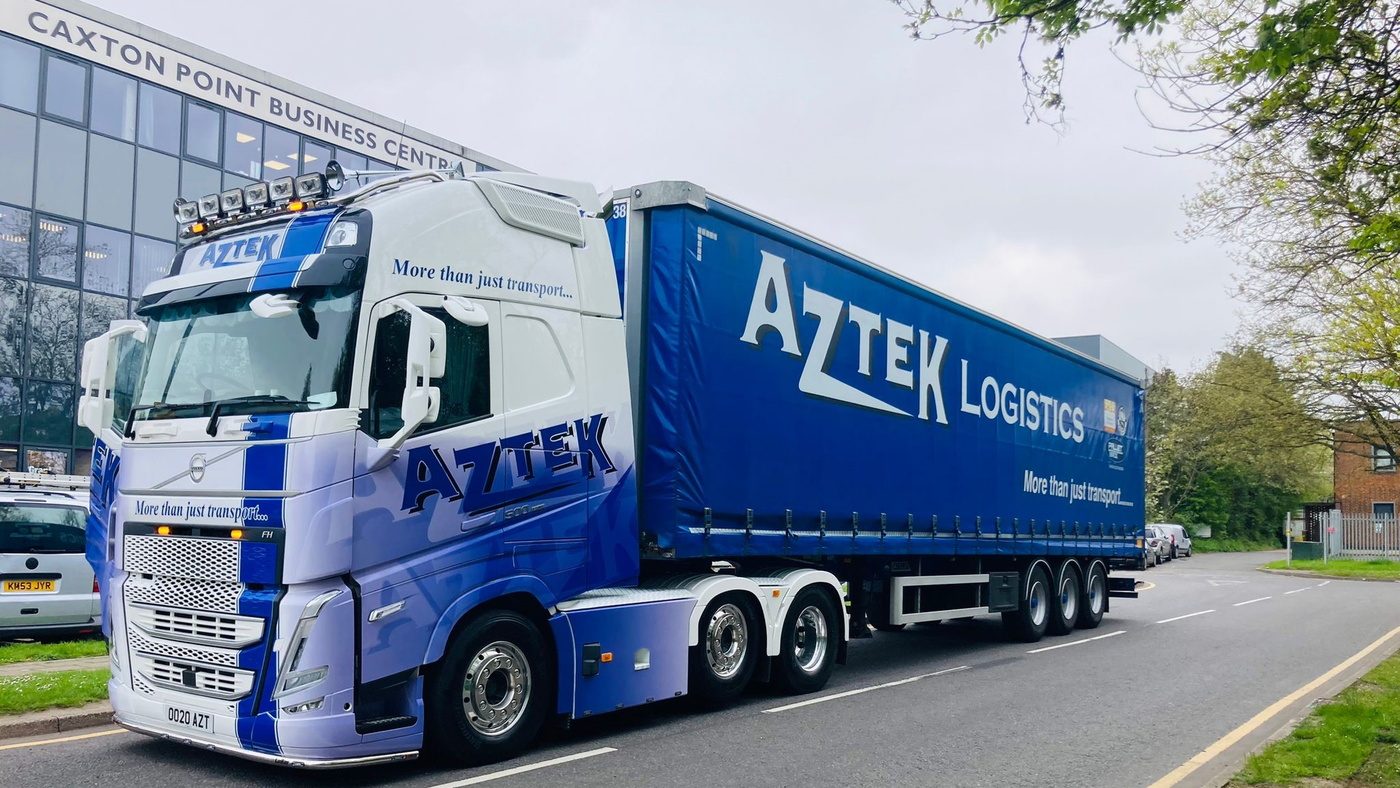 An Aztek Logistics blue and white Volvo tractor unit and curtainside trailer parked outside a business centre