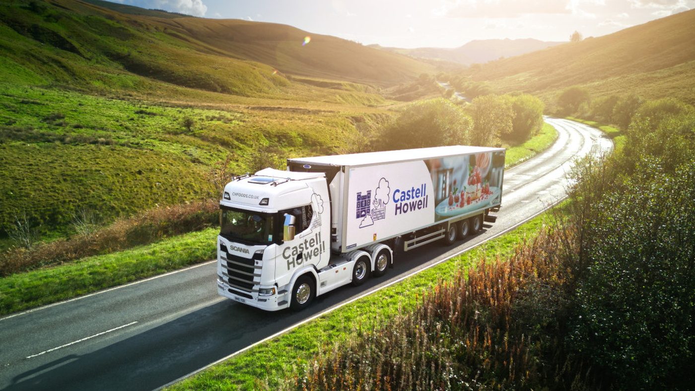 A white Castell Howell refrigerated Scania articulated lorry on a single-carriageway road through green Welsh hills in low sun
