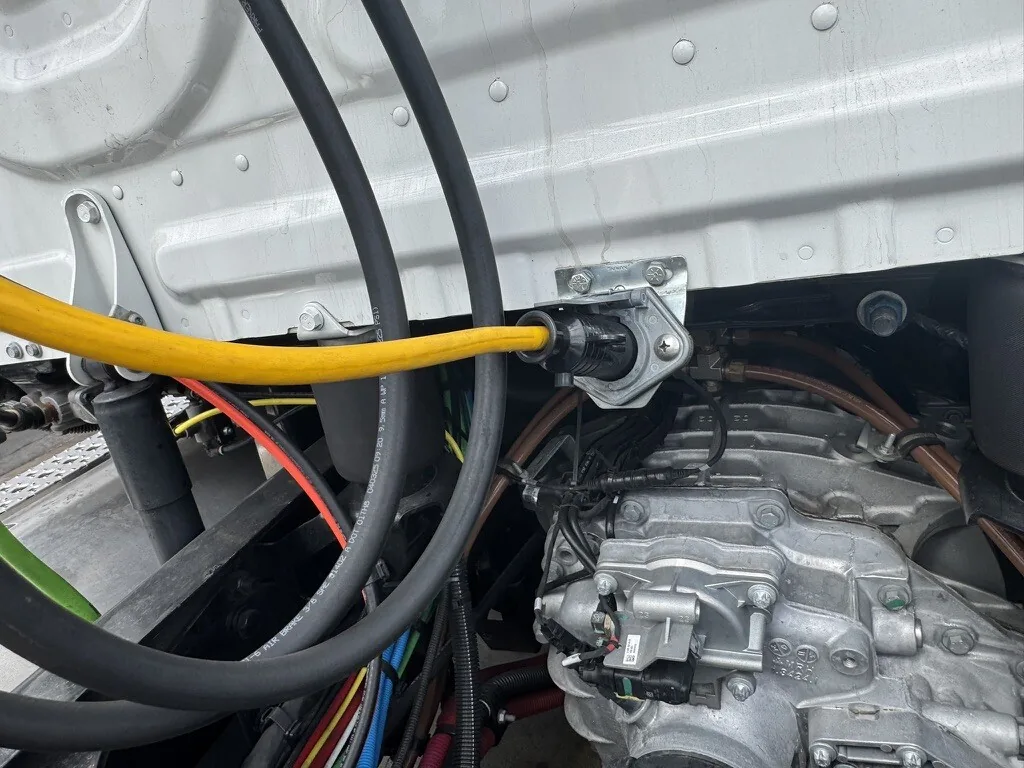 A trailer lead plugged into its socket under the rear of a tractor chassis, beside the air lines