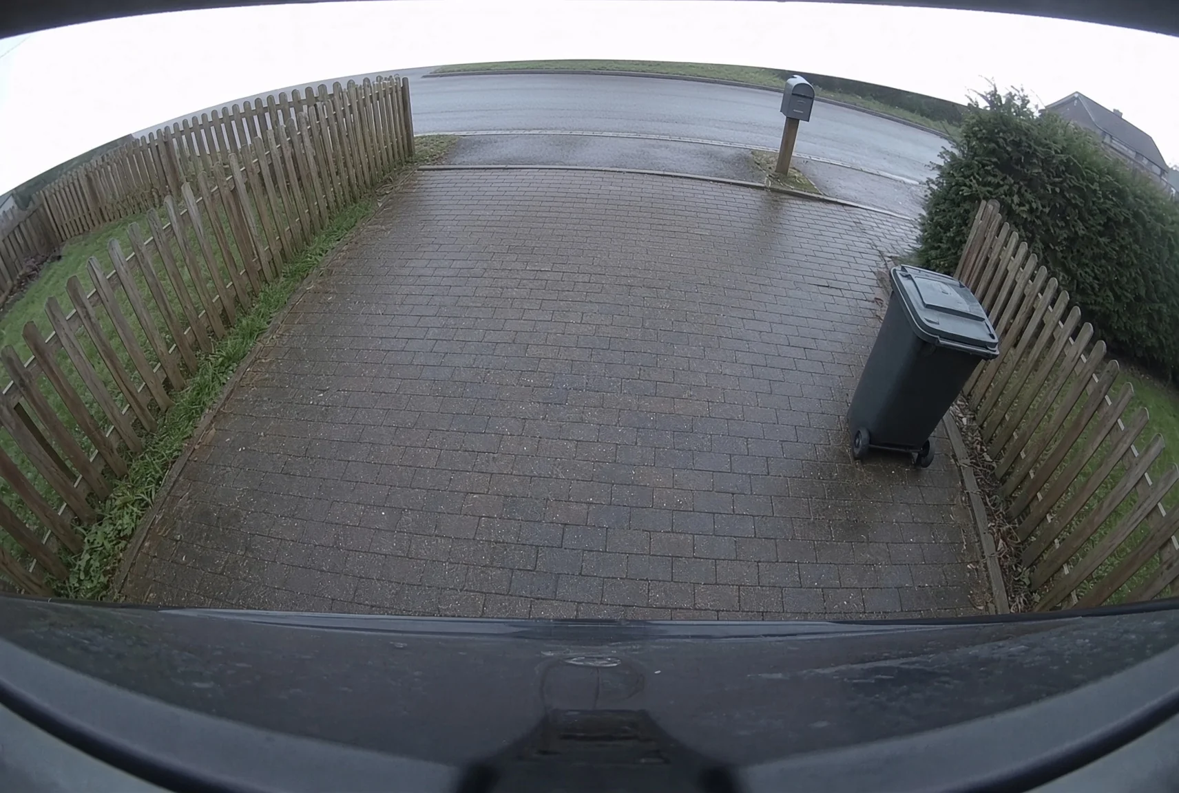 Camera view down a driveway towards the road, with a picket fence to one side, a trash cart and a mailbox at the kerb