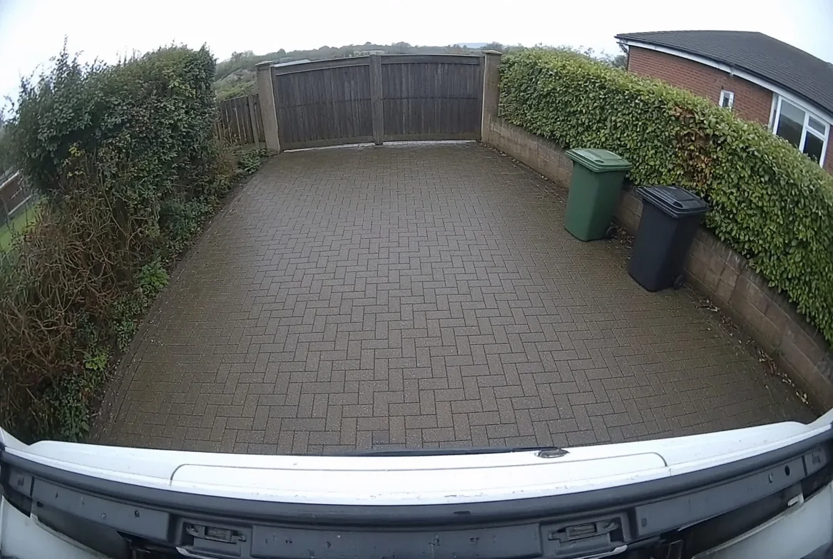 Rear camera view from a van reversing up a block-paved driveway towards a closed timber gate, with wheelie bins against the wall to one side
