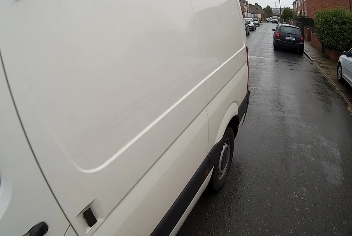 Nearside camera view along a van standing on a residential street, showing the kerb, parked cars ahead and terraced houses beyond